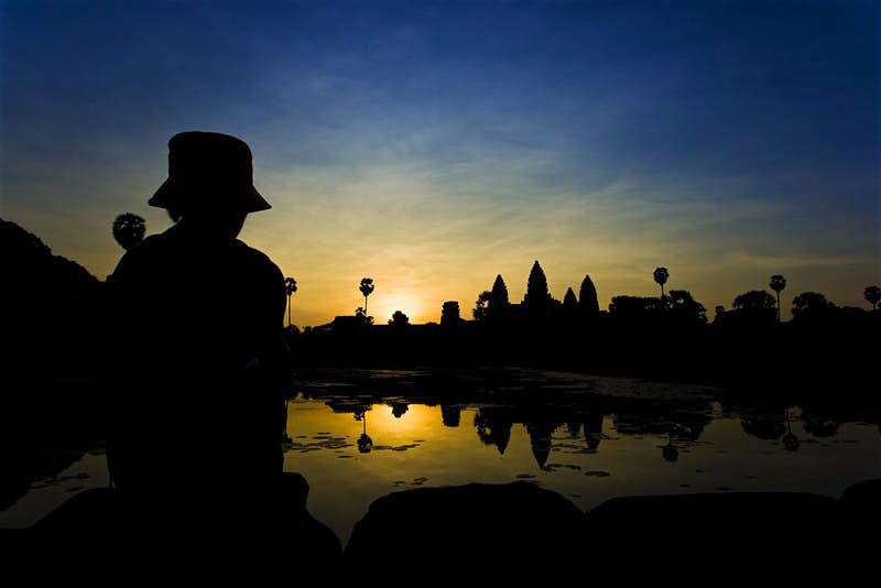 A figure sits in the foreground near the moat watching the sun rise behind the temple of Angkor Wat, which remains in shadow.